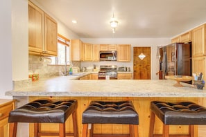 A kitchen with wooden cabinets, a granite countertop, a stainless steel refrigerator, a microwave, a stove, and a sink. There are three leather bar stools. - Game Room + Fireplace! Family Retreat in Pinetop (Pinetop)