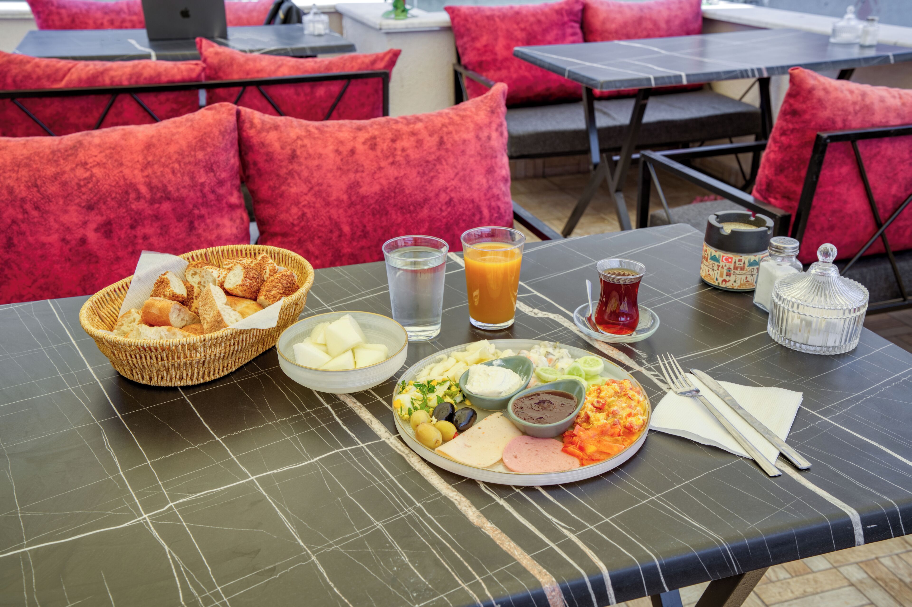 A dining table with a variety of food items, including bread, cheese, olives, and a plate of vegetables. There are also glasses of water and juice, a cup of tea, and condiments on the table.