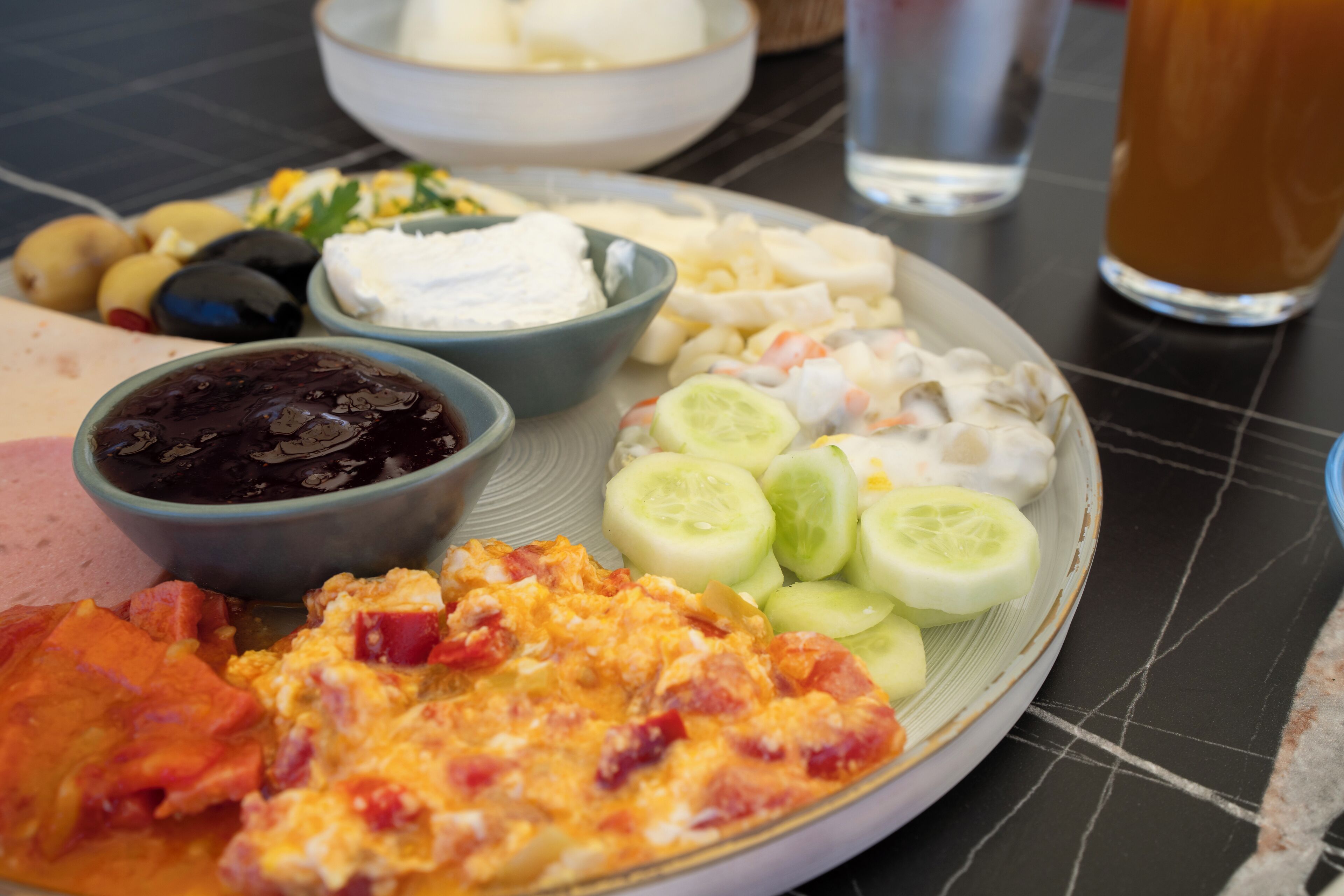 A plate of food with various items including sliced cucumbers, a bowl of black sauce, and a slice of pizza.