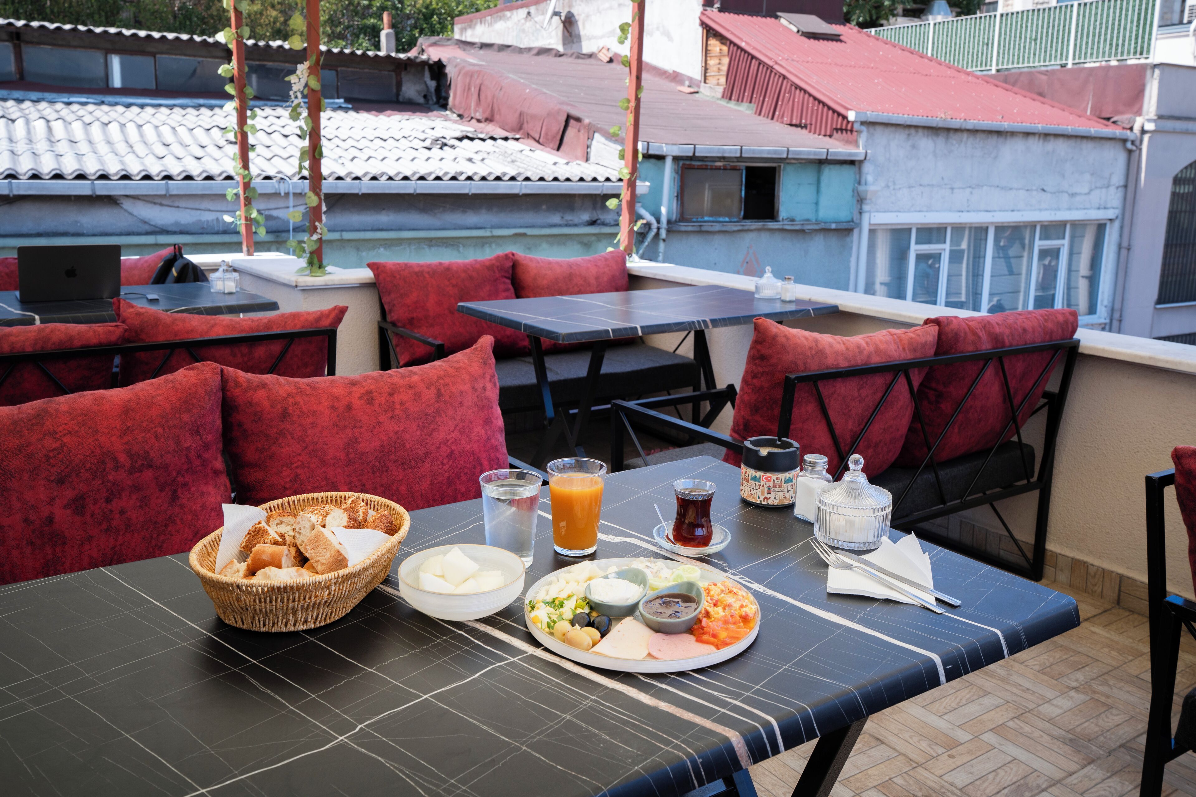 A rooftop dining area with red cushioned seating, a table set with a basket of bread, a plate of food, a glass of orange juice, and condiments.