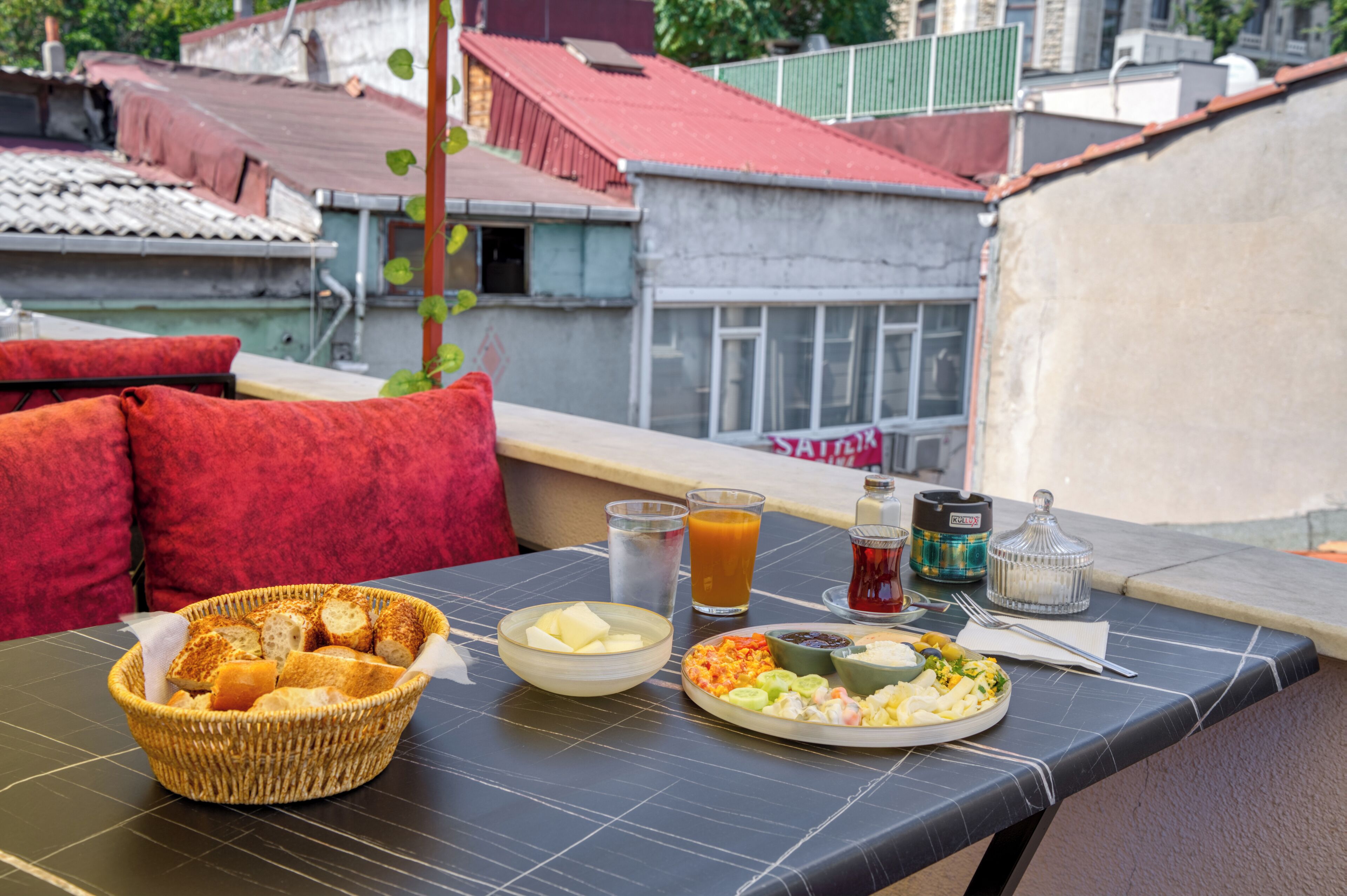 Et morgenbord med ristet brød, smør, frugt og drikkevarer på en balkon med udsigt til bygninger.