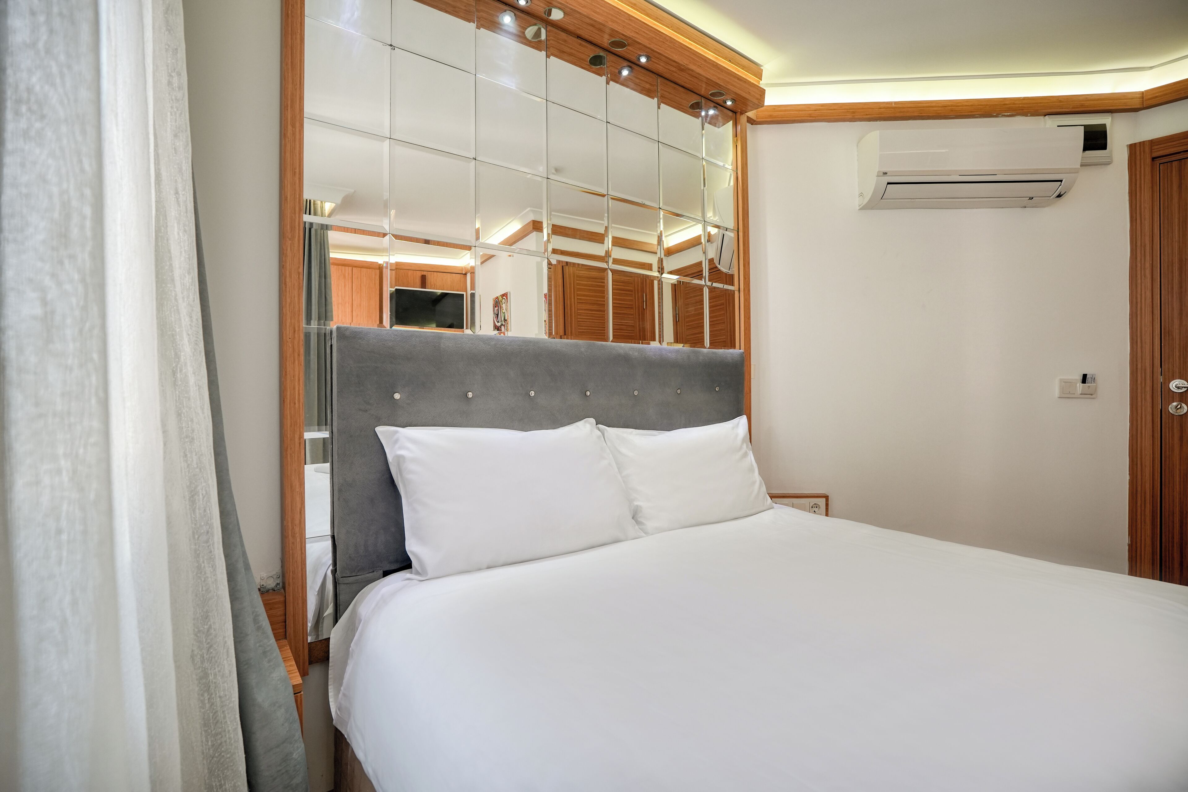 A neatly made bed with white linens in a modern room with a wooden headboard, a large mirror, and an air conditioning unit on the wall.