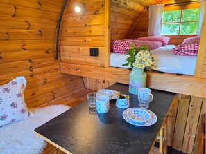 L’intérieur d’un chalet accueillant, avec un décor en bois, un lit recouvert d’une couette à carreaux, une table avec des assiettes et des tasses, et une fenêtre donnant sur la forêt.