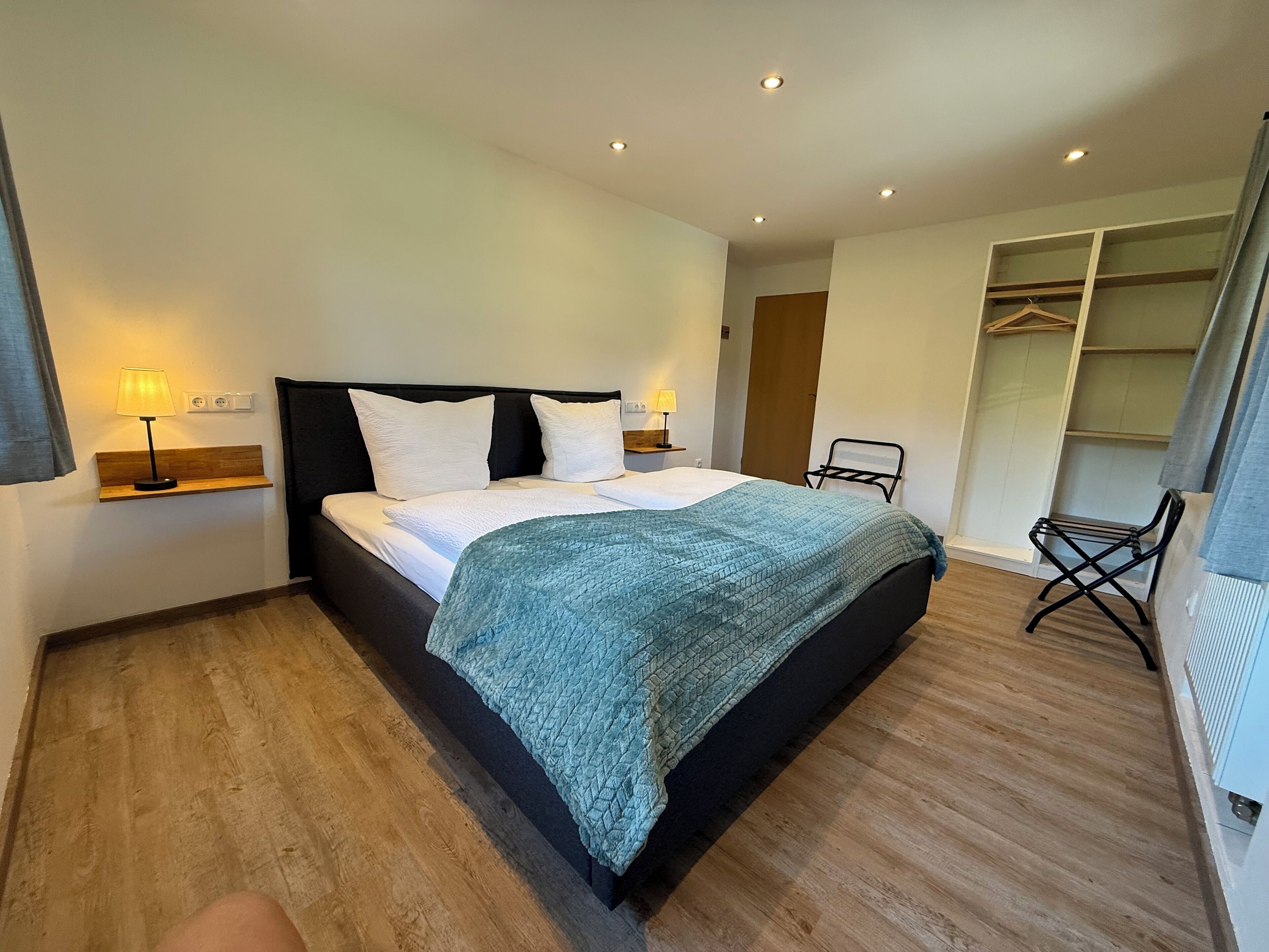 A modern hotel room with a large bed, bedside lamps, a wooden floor, a walk-in closet, and a chair.