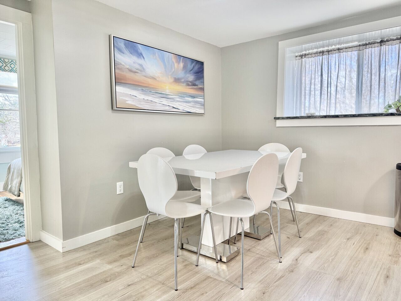 Un coin repas avec une table et des chaises blanches, un tableau au mur et une fenêtre avec des rideaux.