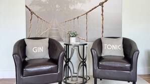Deux fauteuils en cuir ornés de coussins aux motifs « GIN » et « TONIC », une petite table ronde avec une plante, et une balançoire en corde à l’arrière-plan.
