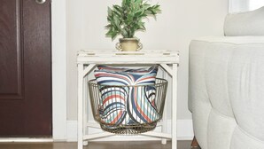 A white side table with a potted plant and a colorful woven basket. - Spacious Stay Near Carowinds Amusement Park | Newly Remodeled | Fenced Yard | Pet & Family Friendly (Pineville)