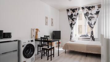 A compact room with a bed, a washing machine, a dining table with chairs, a TV, and a window with patterned curtains.