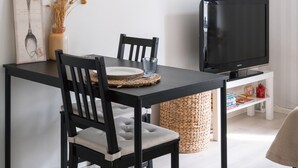 A modern dining area with a black table, two chairs, a woven basket, and a television on a white stand.