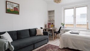 A modern living room with a dark grey sofa, a small dining table, and a large window with a view of the city. - Renovated Top Floor Studio in Kallio (Helsinki)