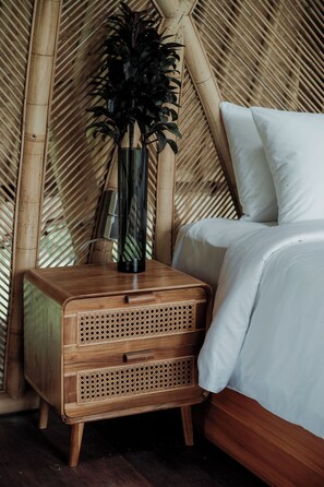 Un lit avec une literie blanche, une table de chevet en bois surmontée d’un panier en osier et un grand vase contenant des feuilles sombres.