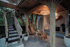 Un escalier avec des rampes en bambou mène à un espace abrité où se trouvent une table et des chaises, le tout entouré de plantes en pot et d’un luminaire suspendu.