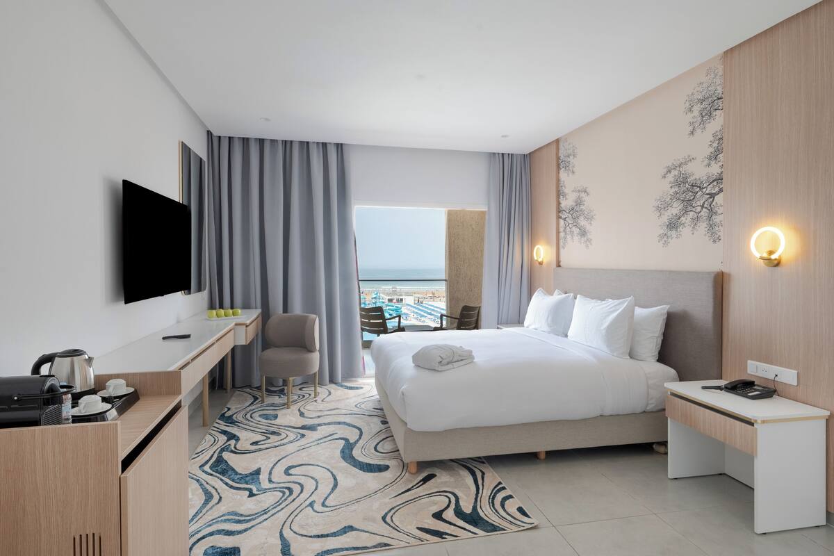 A modern hotel room with a large bed, a desk with a flat-screen TV, a coffee maker, and a view of the beach.