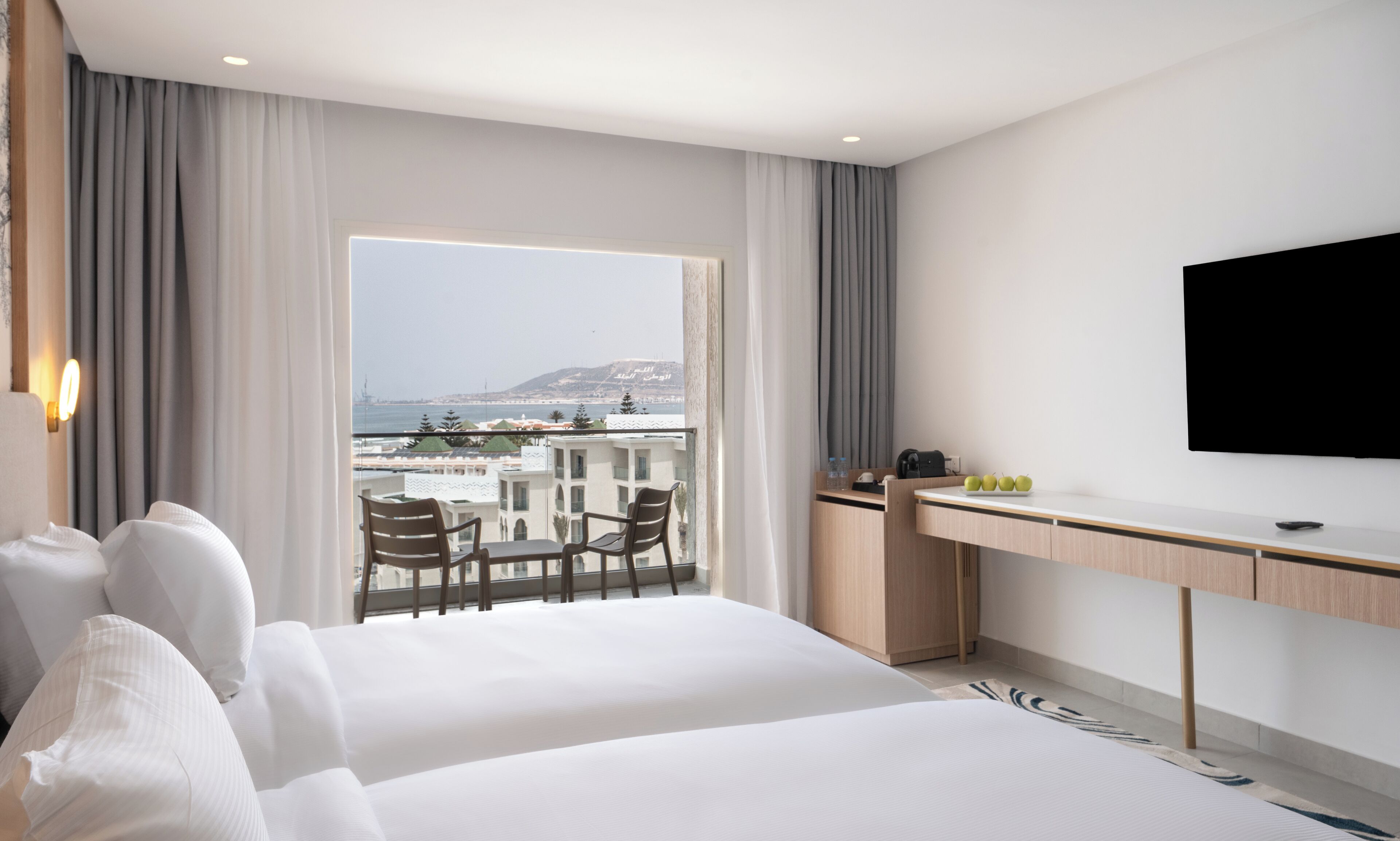 A hotel room with a bed, a desk, a TV, and a balcony with a view of the sea.