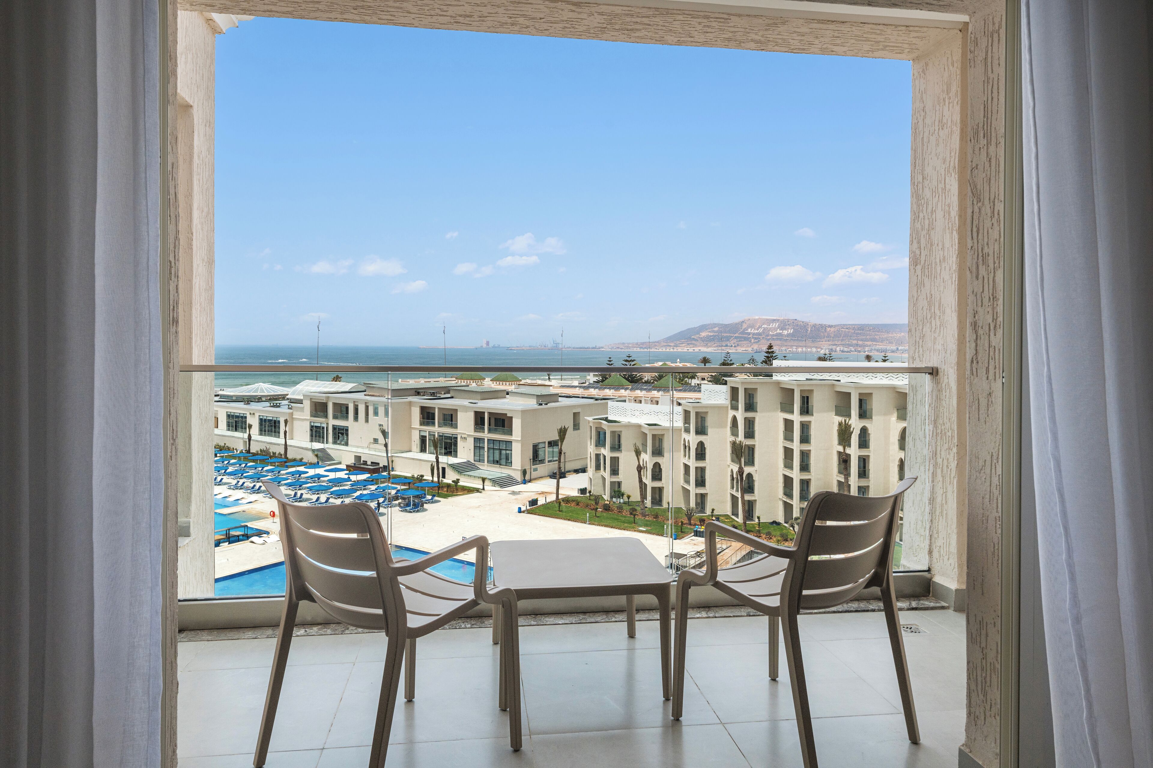A balcony with two chairs and a table, overlooking a pool area with umbrellas and a building complex.