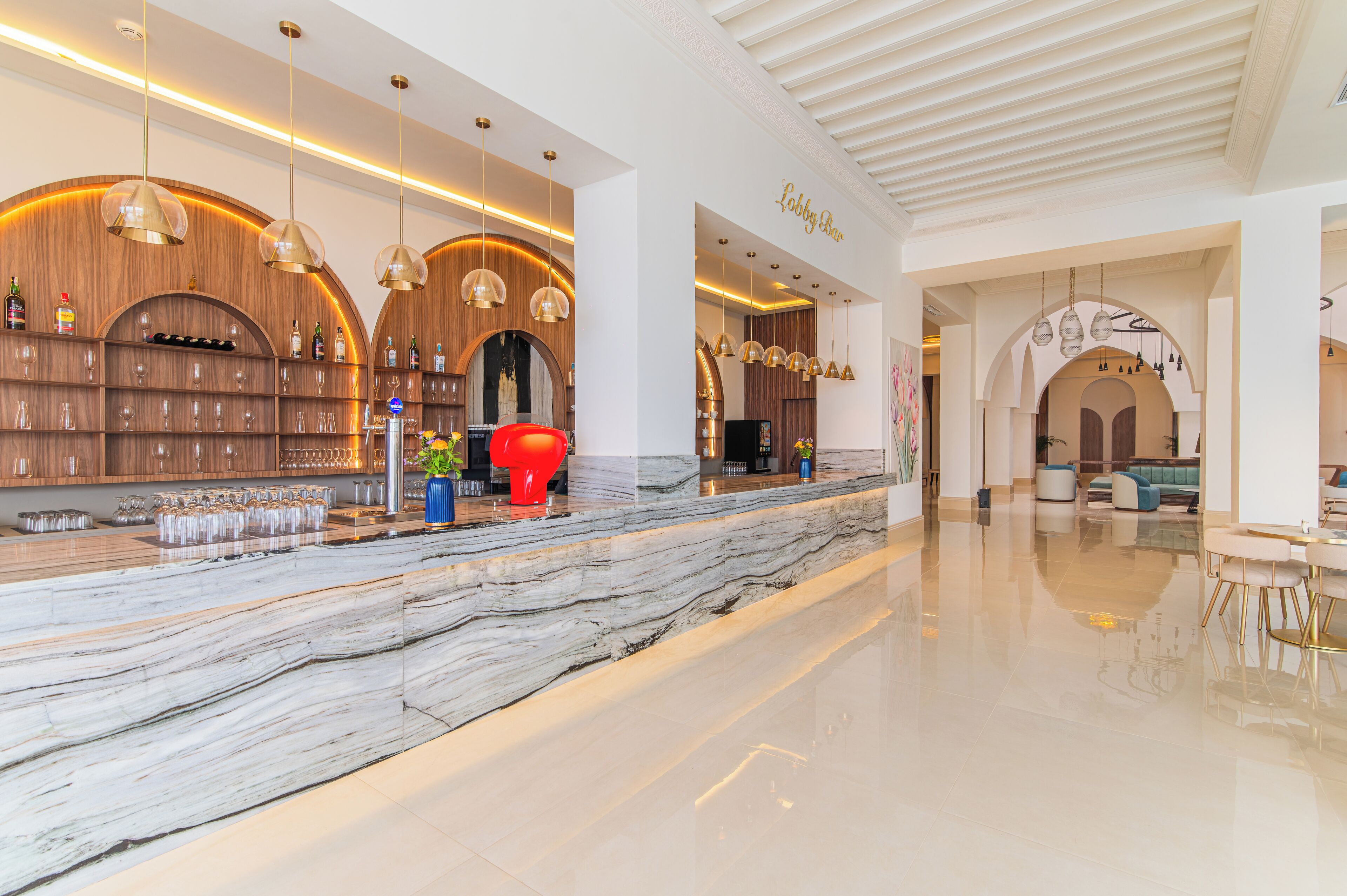 A modern lobby with a marble reception desk, wooden shelving with bottles, and a red sculpture.