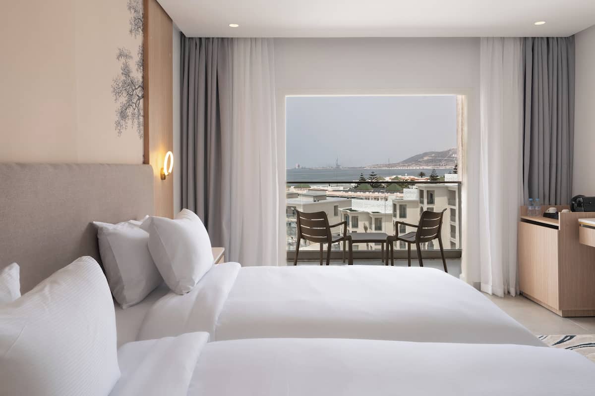 A hotel room with a large bed, a desk, and a balcony with a view of the sea and buildings.