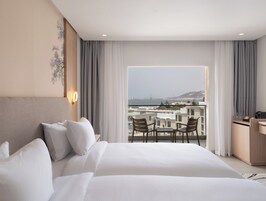 Quarto de hotel com uma cama grande, uma escrivaninha e uma varanda com vista para o mar e edifícios.