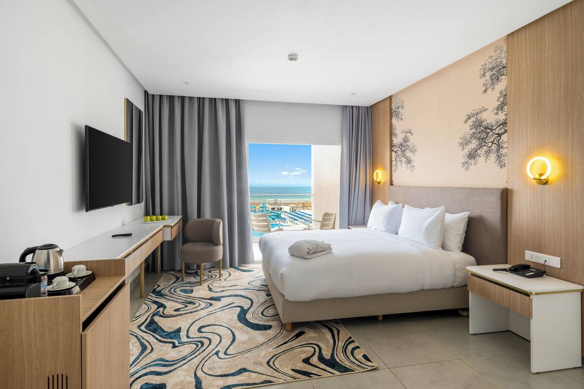 A hotel room with a bed, a desk, a TV, a view of the ocean, and a wall with a decorative tree design.