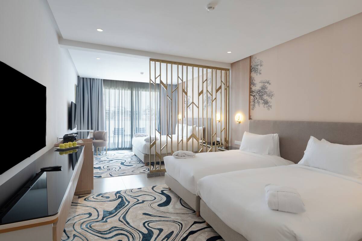 A hotel room with a large bed, a TV, a desk with a computer, and a view of the city through large windows.