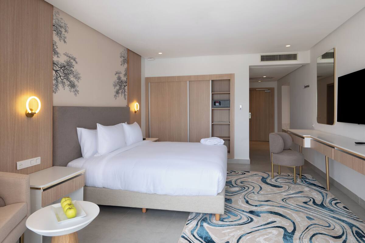 A modern hotel room with a large bed, bedside tables, a desk, a chair, a TV, and a wardrobe.
