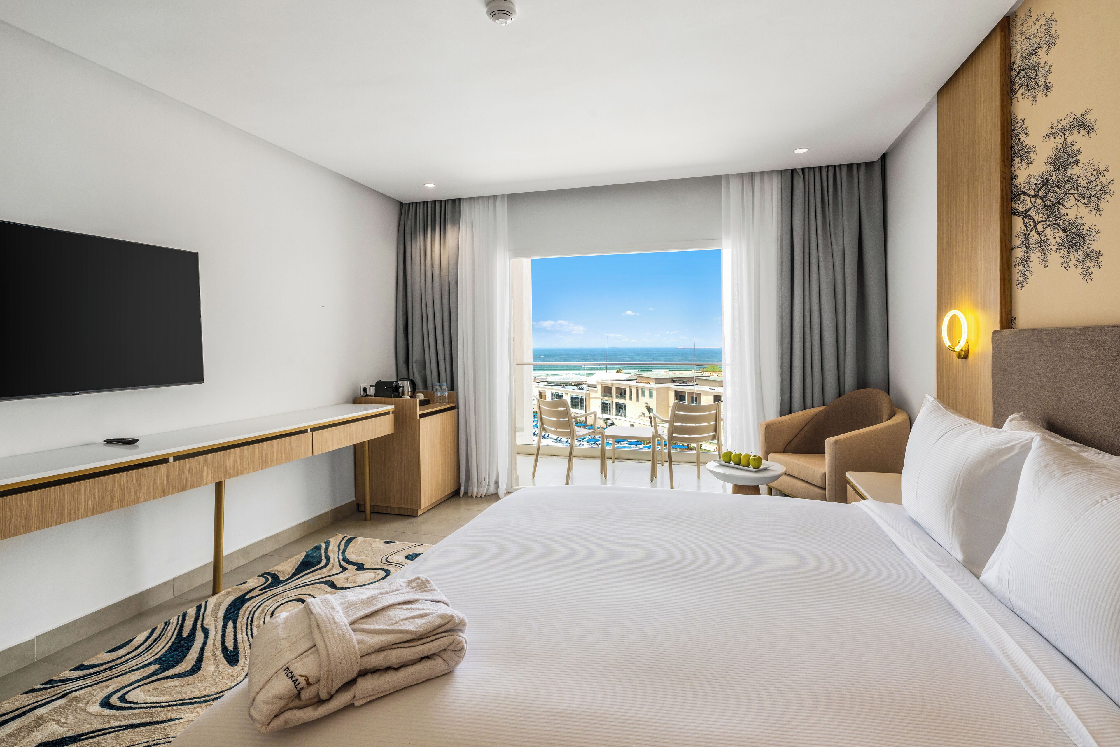 Habitación de hotel con una cama grande, un escritorio, televisión, balcón con vistas al mar y una pequeña mesa con sillas.
