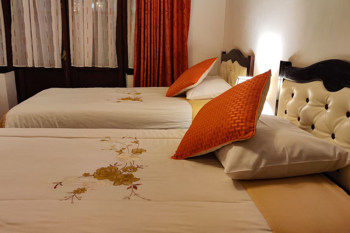 Une chambre d’hôtel avec deux lits, chacun recouvert d’une courtepointe blanche ornée de broderies florales et agrémenté d’oreillers orange.