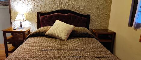 Una cama con colcha estampada y cabecera capitonada, flanqueada por dos mesitas de noche con lámparas y una pequeña mesa.