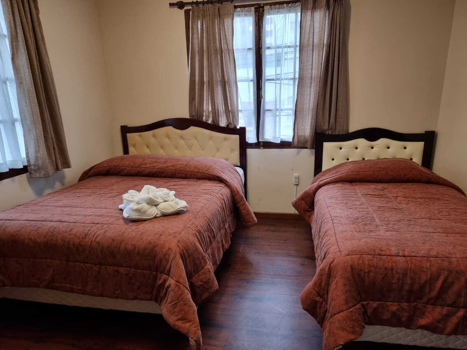 A room with two beds, each with a headboard and a brown bedspread. There is a window with curtains, and a white outlet is visible on the wall.