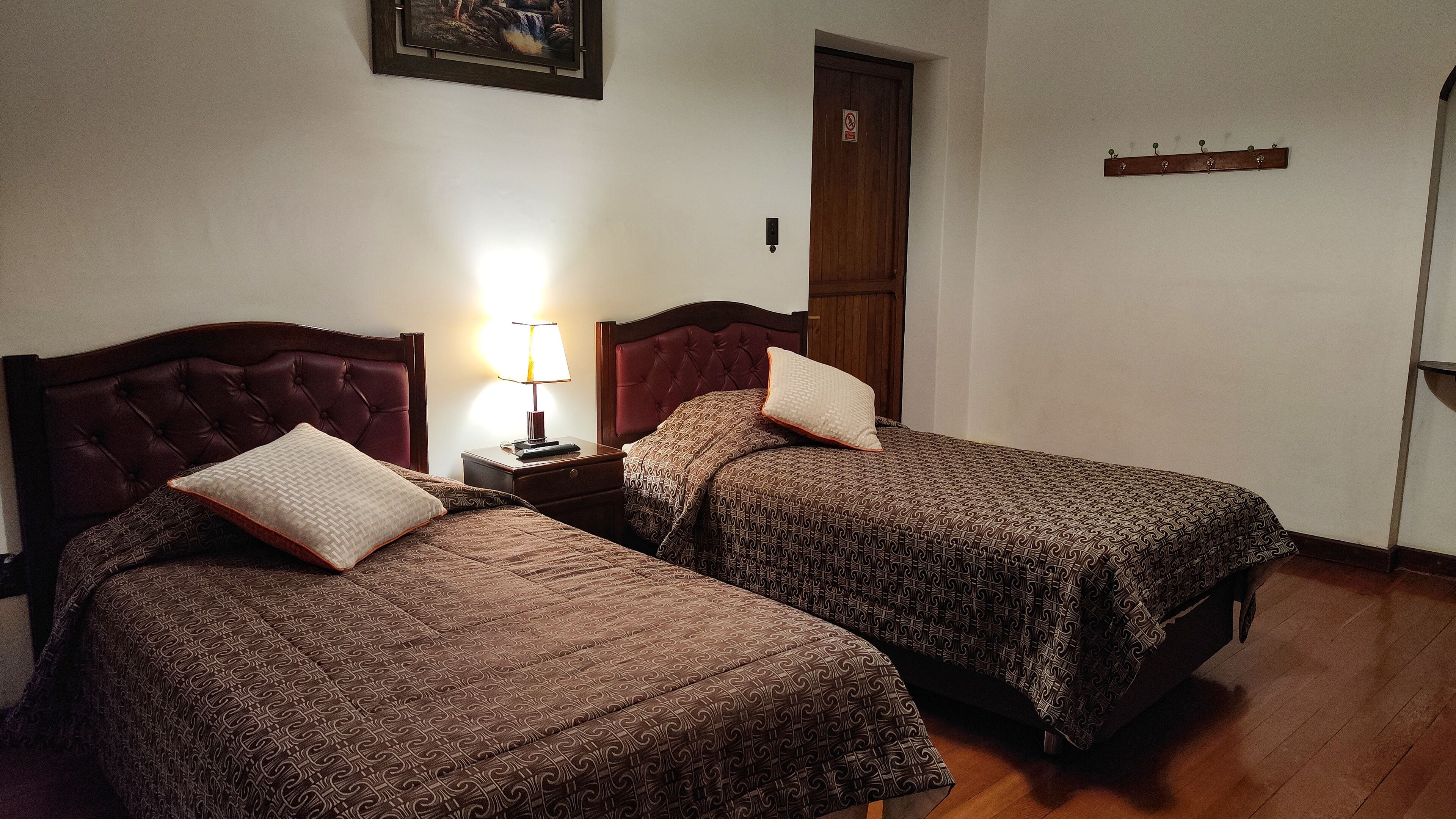 Een hotelkamer met twee bedden, elk voorzien van een beddekleed met een patroon en een kussen. Tussen de bedden staat een nachtkastje met een lamp.