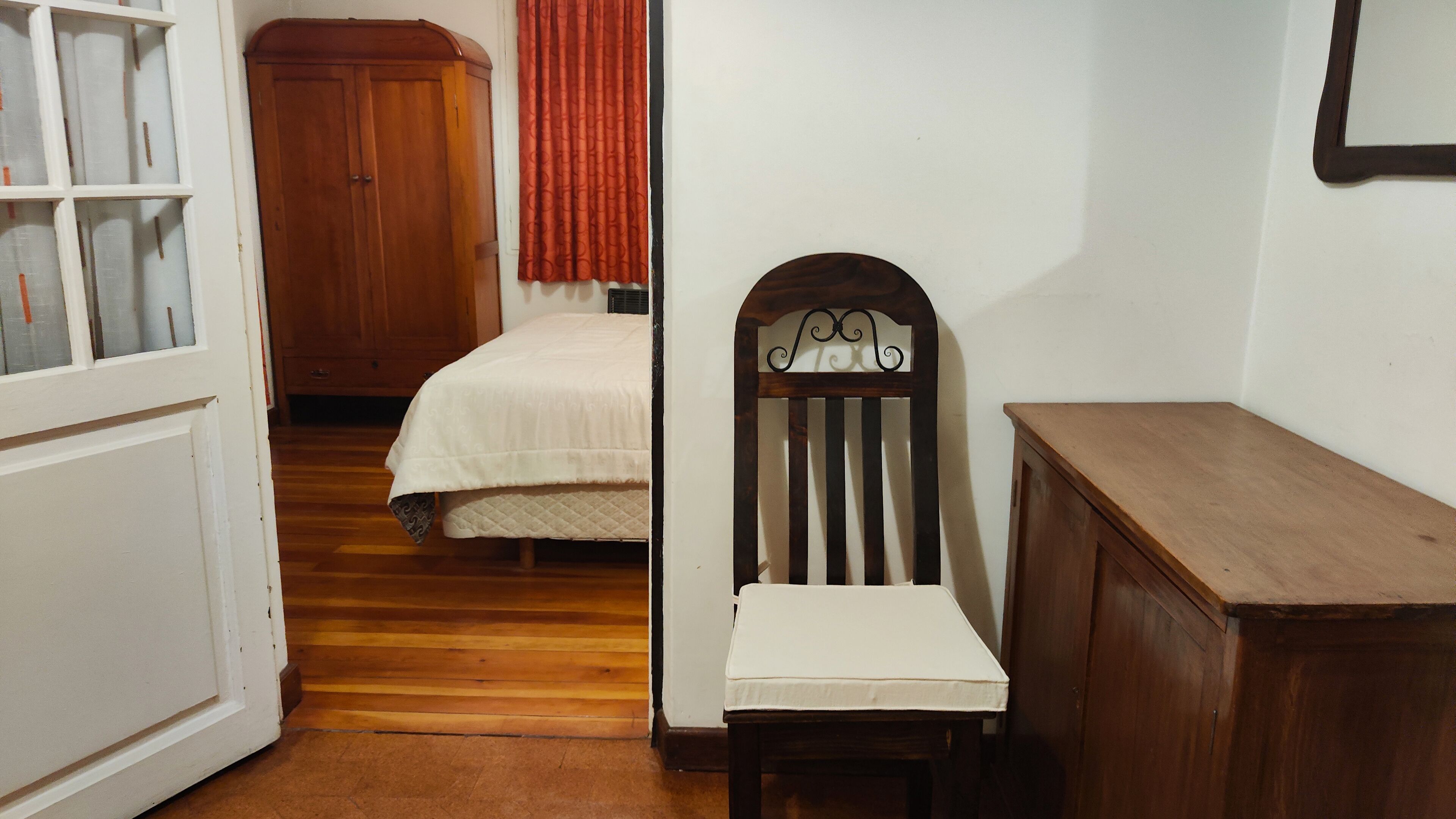 Un couloir étroit avec une armoire en bois, un lit recouvert d’une courtepointe blanche et une chaise avec un coussin blanc.