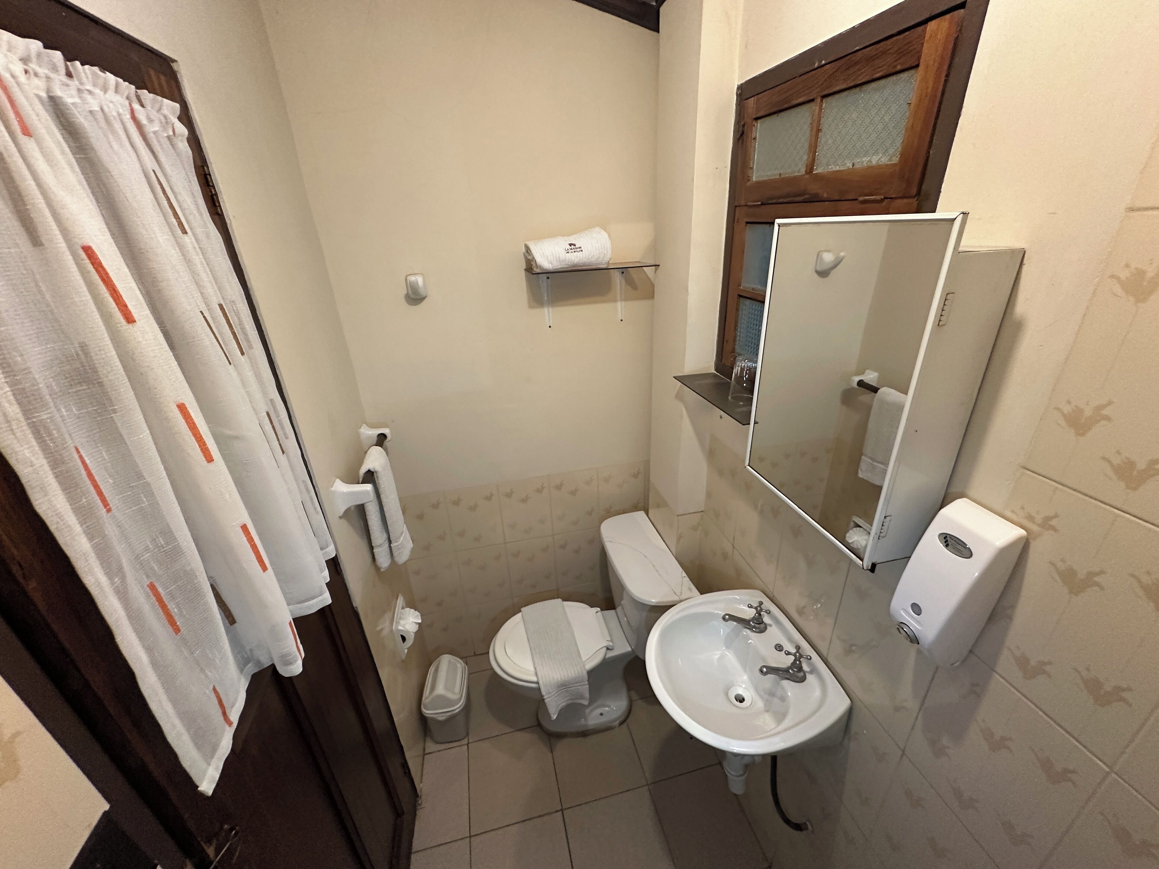 A small bathroom with a toilet, sink, and a mirror. There is a towel rack and a trash bin.