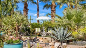 Ein Garten mit Palmen, einem blauen Pflanzgefäß und einer Terrasse mit Stühlen und einem Tisch.