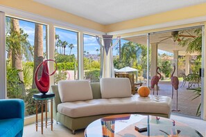 A modern living room with a curved sofa, a glass coffee table, and a view of a garden with palm trees and a pool. - Fun in the Sun! South Palm Desert Gem! (Palm Desert)