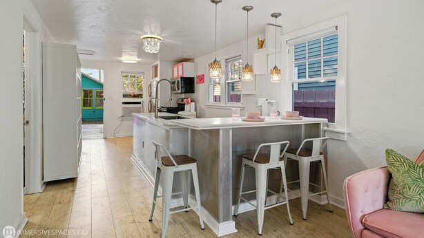 A kitchen with a central island, bar stools, pendant lights, and a view of a blue house through the window. - Summer Family Fun, Pet Friendly, Walk Downtown, Close to Beach, Fenced Courtyard (Lake Worth Beach)