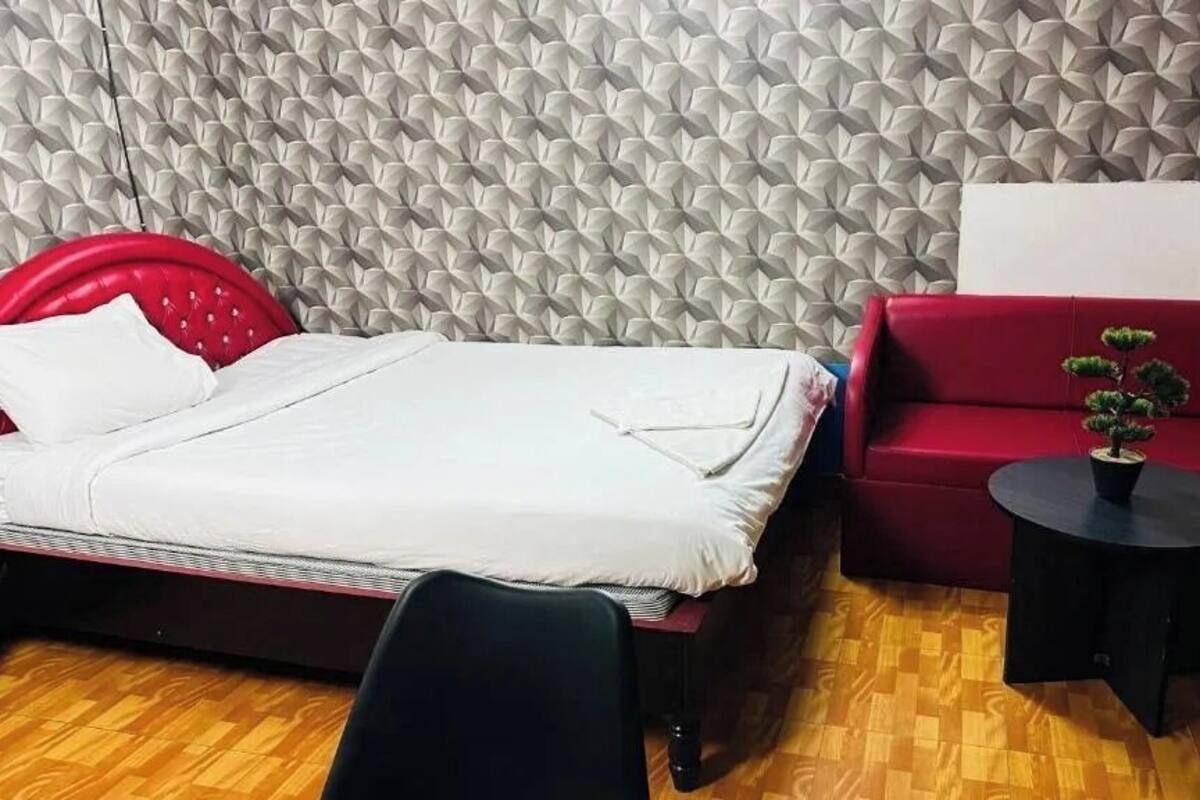 A room with a bed, a red sofa, a small round table with a plant, and a textured wall.