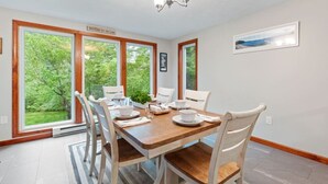 Comedor con mesa de madera, sillas blancas y vista a la vegetación a través de amplias ventanas.
