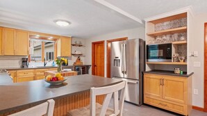 Una cocina moderna con armarios de madera, un refrigerador de acero inoxidable, un microondas y un comedor con un tazón de fruta y una silla.