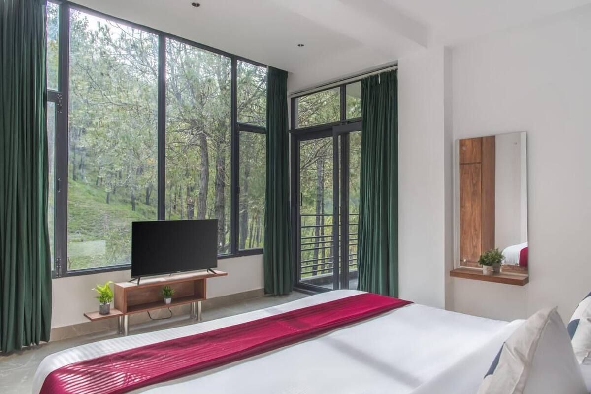 A bedroom with a large bed, a TV on a wooden stand, and a view of trees through large windows.