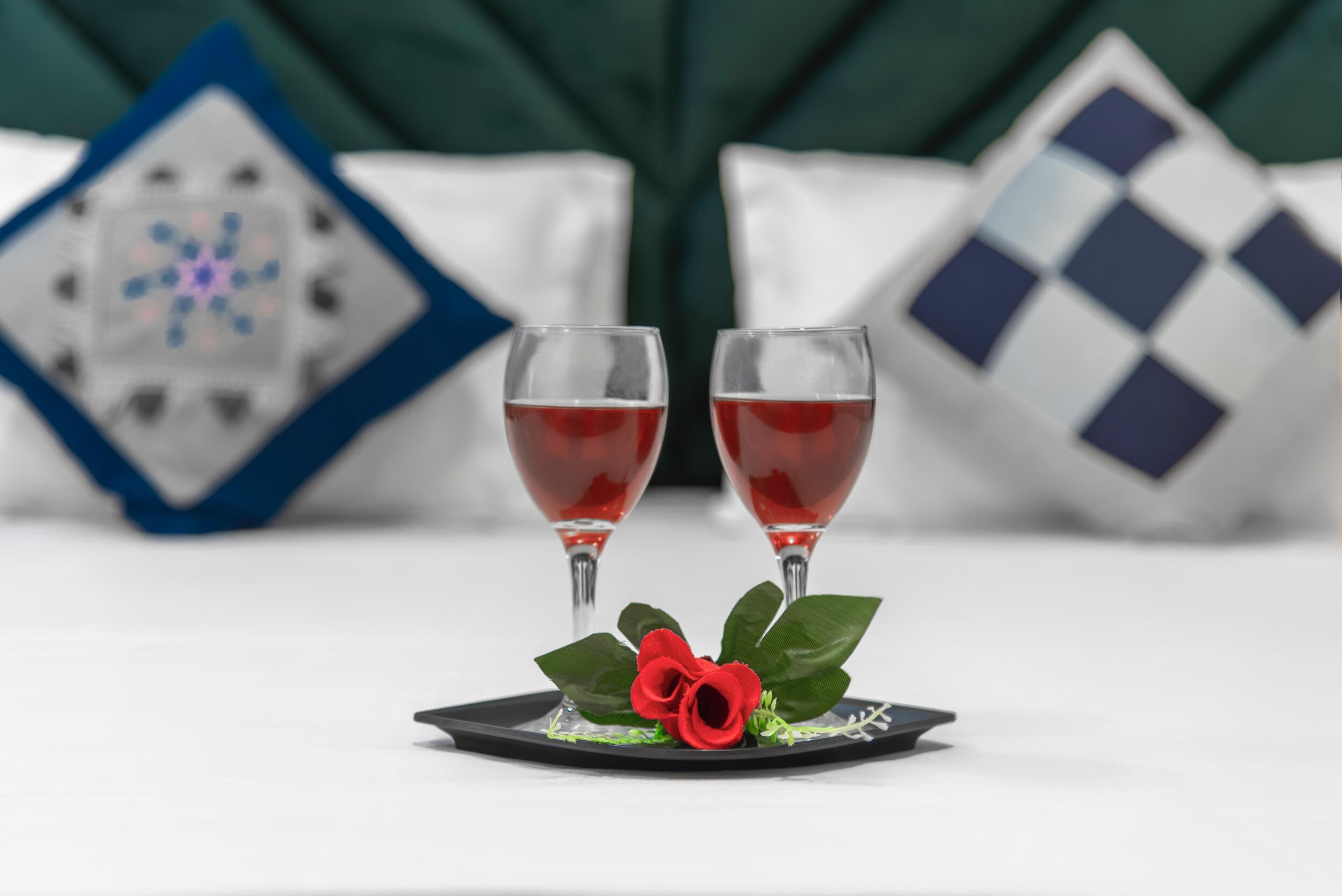 Two glasses of red beverage on a tray with a red rose decoration, set against a background with decorative pillows.