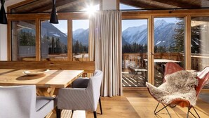 A dining area with a wooden table and chairs, a view of mountains through large windows, and a cozy chair with a fur throw.