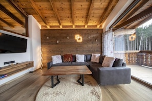 Un salon moderne avec un plafond en bois, un grand téléviseur à écran plat, un canapé d’angle gris et une table basse en bois.