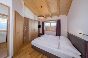 Une chambre avec un plafond en bois, un lit recouvert de draps blancs, un téléviseur fixé au mur et une fenêtre avec des rideaux.