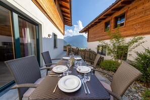 Un espace de restauration en plein air, avec des chaises en osier et une table dressée pour quatre personnes, entouré de bâtiments en bois et offrant une vue sur la montagne.