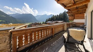 A wooden balcony with a view of mountains, stone walls, and a rustic outdoor seating area.