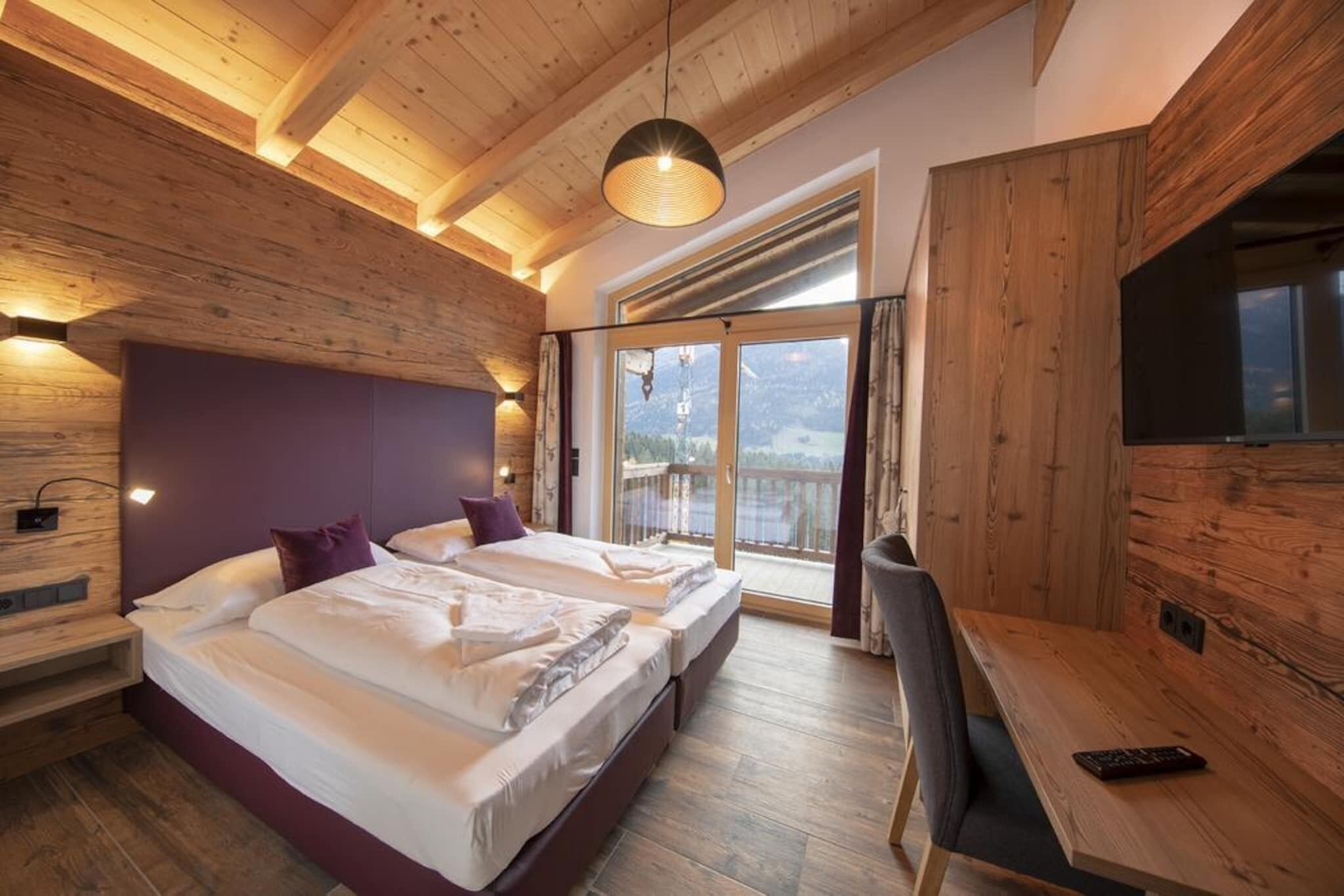 A bedroom with two beds, a desk, a TV, and a balcony with a mountain view.