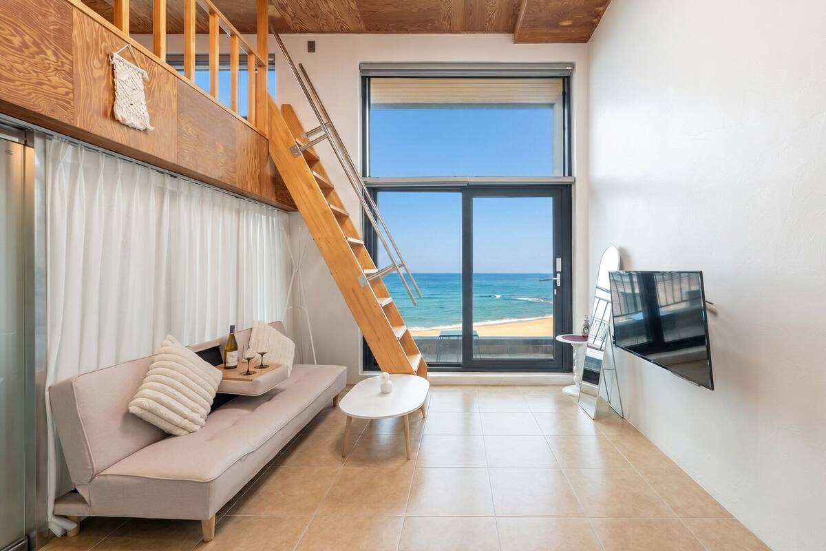 A modern living room with a sofa, a coffee table, a TV mounted on the wall, a wooden staircase, and a large window with a beach view.