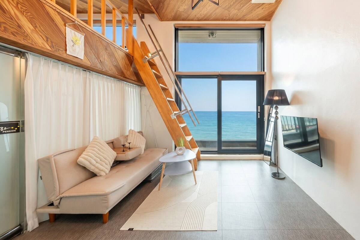 A modern living room with a sofa, a coffee table, a TV, and a view of the sea through a large window.