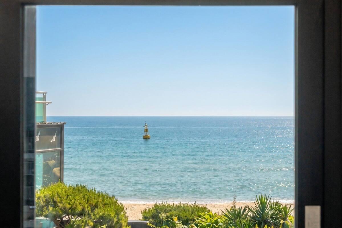 A view of the sea from a window, with a buoy in the water and a beach in the foreground.