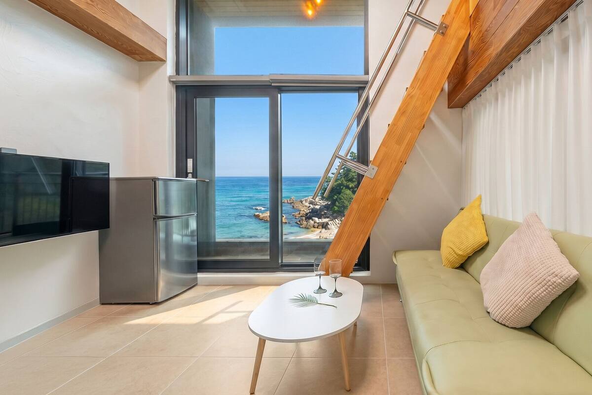A modern living room with a sofa, a small table, a TV, and a large window offering a view of the sea.